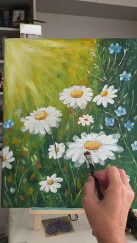 A hand holding a paintbrush is adding white and orange paint to a daisy flower in a painting. The painting depicts a field of daisies with a bright yellow sunlit background. Other small blue flowers are also visible in the painting. The painting is on an easel.