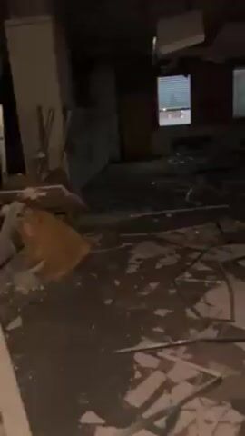The video presents a very dark and dilapidated indoor space, potentially an abandoned building. The room is filled with debris scattered across the floor. There appear to be windows along one side of the room, letting in dim natural light. There are also white vertical pillars or partitions within the room. The general atmosphere is eerie and decaying.