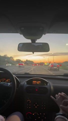 The video shows the view from inside a car driving on a highway at dusk. The sky is a mix of blue and orange. Several other cars are visible on the road. The dashboard of the car is in focus, showing the speedometer, radio, and other controls. The time displayed on the radio is 20:06. Two hands are visible in the lower right corner of the frame, holding each other. The windshield is slightly dirty. Streetlights line the highway.