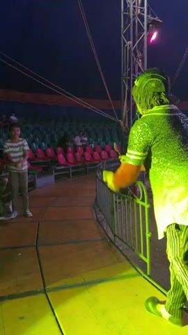 A male clown in green clothing, including a baseball cap, a sequin-covered shirt, and striped pants, is on a yellow-lit stage inside a circus tent. He holds a fork in his hand and walks back and forth along a metal barrier surrounding the stage. A young boy in a striped shirt stands to his left, and there are a few people seated in red and green chairs in the background. A tall metal structure with a spotlight is on the right side of the frame.