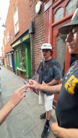 The video shows a narrow outdoor street scene involving three or four young men. The environment consists of buildings with varied painted storefronts and brick facades. The street is paved with cobblestones. One man, wearing a black t-shirt with a yellow design and a dark hat, appears to be handing something to another man, obscured from the view. A third man, wearing a gray t-shirt and a white cap, is standing nearby. Subsequently, the focus shifts to a different man wearing a red t-shirt who is smoking. In the background, the man who was previously walking towards the camera, now stands still, holding a green bottle.