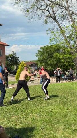 Two shirtless men are wrestling on a grassy field, encircled by a gathering of spectators. One man is wearing black pants with white stripes on the side, while the other is wearing solid black pants. They are grappling with each other, and at one point, one lifts the other off the ground. Around the wrestling area, people stand and watch, some sitting on benches under trees. In the background, there is a building, a small evergreen tree, and a few utility poles. The sky is blue with scattered clouds, suggesting a daytime outdoor setting. A man in a blue shirt and black pants stands on the sidelines, attentively watching the wrestling match.