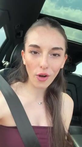 The media file is a close-up, slightly shaky video of a young woman with dark brown hair and prominent freckles. She is seated in a car, indicated by the presence of a seatbelt and the interior of the vehicle, including a sunroof and dark leather seats. The woman is wearing a dark red strapless top and a delicate necklace with a small, colorful pendant. Her fingernails are painted a pale color, and she wears a bracelet on her right wrist.

The woman displays a range of emotions, primarily happiness and playfulness. She smiles broadly, laughs, and makes exaggerated facial expressions, including puckering her lips and gesturing with her hands. At one point, she touches her chin thoughtfully with her finger. The lighting suggests it is daytime, likely with bright, possibly sunny weather, as evidenced by the illumination and the view through the sunroof of a cloudy sky. The scene appears to be outdoors, possibly on a road trip or traveling. There is no visible text in the media file. The setting is implied to be Chokoba, Bulgaria, as per the provided location context.