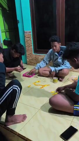 Four young men are sitting on a tiled floor playing dominoes. The man on the far left is wearing a black t-shirt and black pants with white stripes. He is holding a smartphone and looking at it. The man next to him, wearing a grey hoodie and shorts, is also looking at the phone. The third man, wearing a light blue hoodie and shorts, is looking at the dominoes on the floor. The fourth man on the right is wearing a blue and yellow striped t-shirt and blue shorts. He is also looking at the dominoes. The dominoes are arranged in a line on the floor. There is a pink container filled with dominoes and a small yellow cup on the floor. The background shows a teal wall and a window with reflections. The lighting is dim, suggesting it is evening or nighttime.