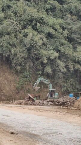 The video shows an excavator in operation at a construction site next to a densely vegetated hillside. The excavator is pale green and is positioned on a dirt road, using its bucket to scoop rocks and deposit them into a nearby sifter, which appears to be attached to a wheeled trailer.  A pile of rocks is also present on the ground next to the excavator.  In the background, a steep hillside is covered with trees and bamboo. The scene suggests a road construction or maintenance project in a remote, possibly mountainous area. No people are visible in the video.