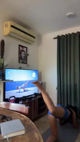 The video shows a woman in blue shorts and a black top doing leg exercises while watching a video on TV, which appears to be a workout tutorial. The room has green curtains, a Che Guevara poster, and a wooden TV stand. Later, an older woman in a floral dress stands near a couch and a staircase. The camera then focuses on a plate of watermelon slices on a glass table. Finally, a young woman in a gray t-shirt takes a selfie, making a peace sign gesture.