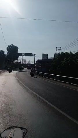 The video shows a first-person perspective of riding a motorcycle on a road. The camera is pointed forward, capturing the road ahead and the surrounding environment. The sky is clear and bright, with the sun visible on the left side of the frame, creating lens flare and casting shadows. The road appears to be asphalt and is in good condition, with a white line marking the edge on the right side.

Several vehicles are on the road, including cars and motorcycles. On the right side of the road, there is a barrier or fence with vegetation behind it. Overhead, there are power lines and a large sign structure with directional signs. In the distance, there are buildings and trees. The video captures the movement of other motorcyclists in front and to the side, as well as other vehicles in the background. The overall scene depicts a sunny day on a moderately busy road.