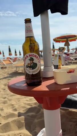The video shows a bottle of Birra Moretti beer on a small orange table on a sandy beach. In the background, beach umbrellas and lounge chairs are visible under a clear blue sky with some clouds. The bottle is the primary focus, with its label clearly readable. To the right of the beer bottle, there is a white plastic container holding some red items, possibly food. The scene appears to be a relaxed, sunny day at the beach.