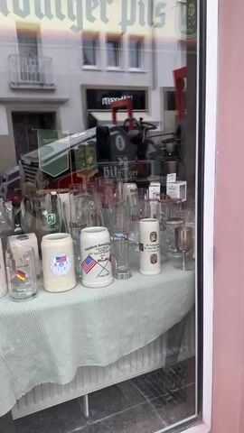 The video shows a storefront window display. In the foreground, on the left, is a table covered with a green and white checkered cloth. On the table are several beer glasses of various shapes and sizes, some with logos. There is also a white beer stein with a logo. A framed advertisement featuring a man's face and the words "Bitburger Pils" is on the bottom left of the window. To the right of the table, a life-sized statue of a man in a green uniform is holding a large beer bottle. The man has blond hair and is smiling. Behind the statue, more items are visible, including a vintage camera, boxes, and a radiator. Reflections of the street and buildings across the road are visible in the window. A woman is visible in the reflection on the right side of the window, taking a photo with her phone.