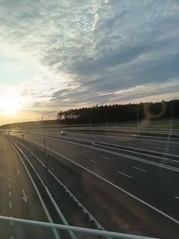 A drone shot from above a highway at sunset. The highway has multiple lanes, and there are a few cars visible. The sky is filled with clouds, and the sun is setting on the horizon, casting a warm glow over the scene. A line of trees can be seen in the distance. The camera pans slowly to the right, showing more of the highway and the surrounding landscape. The video ends with a shot of the highway curving to the right.