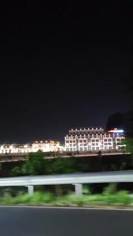 The video shows a nighttime view from a moving vehicle, likely a car, looking towards a building complex. The sky is dark, indicating it is night. The building in the foreground is large and illuminated with numerous lights, giving it a modern appearance. It has multiple floors and windows. To the left of this main building, there is another building, also illuminated, with a lighter color and a different architectural style. Between the viewer and the buildings, there is a raised road divider or barrier, and some greenery is visible behind it. The vehicle's headlights create streaks of light in the lower part of the frame, suggesting motion. The overall scene conveys a sense of urban or suburban nighttime landscape.