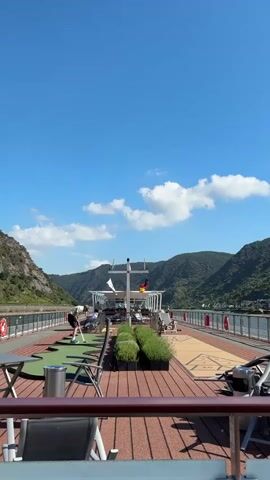 The video shows a wide shot of the deck of a cruise ship on a sunny day. The sky is blue with scattered white clouds. On the deck, there is a putting green area where a person in a white t-shirt and shorts is practicing golf. There are also tables and chairs, planters with green plants, and a shuffleboard court. In the background, a German flag is visible on a flagpole. The ship is sailing through a scenic river valley with lush green hills on either side. The water of the river is visible on the left and right of the ship. The railing of the ship is in the foreground.