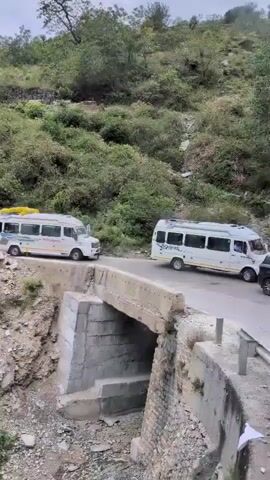 The video shows a winding road in a mountainous, forested region. There are several vehicles, including white vans and cars, stopped on the road, suggesting a traffic jam or blockage. The road is narrow and curves along the side of a steep, rocky hill covered in green vegetation. A small concrete bridge is visible over a gully. The sky is overcast, indicating possibly inclement weather. The overall impression is a remote, rural landscape experiencing a temporary halt in traffic.