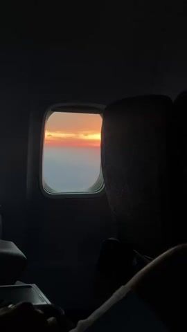 A first-person view from an airplane window shows a sunset. The window is framed by the dark interior of the plane, with the edge of a seat visible on the right. The sky outside is a vibrant mix of orange, pink, and yellow, with clouds streaked across the horizon. Below the sky, a hazy landscape can be seen, possibly land or ocean. The lighting is dim, characteristic of an evening flight.