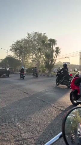 A vertical, slightly shaky video shows a view from a moving motorcycle on an asphalt road. The sun is low in the sky, casting a warm light and creating a hazy atmosphere. Several other motorcycles, ridden by people wearing helmets and casual clothing, are also on the road. In the background, there are trees, streetlights, and a sidewalk with some parked cars. The road appears to have some cracks and wear. The perspective is from the rider of the motorcycle, with a rearview mirror visible in the lower right corner of the frame. The video captures a moment of daily commuting in what appears to be a tropical or subtropical environment.