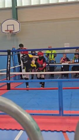 The video shows two young people sparring in a boxing ring. Both boxers are wearing protective headgear, black shin guards, and dark colored shorts. One is wearing black and yellow, the other red and black protective gear. They throw punches and kicks at each other in the ring. Several people stand outside the boxing ring, including adults. Some are watching the action, and some are sitting at a table. The ring is blue and red. There is a basketball hoop on the wall. The camera angle is from outside the ring.