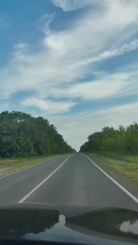 The video shows a view from inside a vehicle driving on a paved road. The road is bordered by lush green trees on both sides, creating a tunnel-like effect. The sky above is a vibrant blue, with scattered white clouds. The road appears to be mostly empty, with only a few distant vehicles visible. The sun is shining, casting reflections on the road surface and the vehicle's dashboard. The overall scene suggests a pleasant drive through a natural landscape.