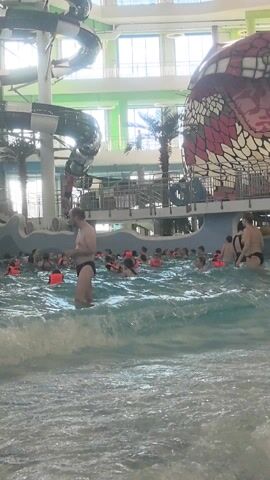 Here is a description of the video:
The video shows a wave pool indoors. There are people in the water, some wearing life jackets. A woman in a black swimsuit stands in the foreground, holding a baby. There are water slides and decorative palm trees visible in the background.