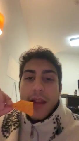 A young man with dark curly hair and a mustache eats an orange tortilla chip in a white room. He is wearing a light-colored fleece jacket. A desk and computer monitor are partially visible in the background. An orange-colored light fixture is visible in the upper-left corner of the frame.