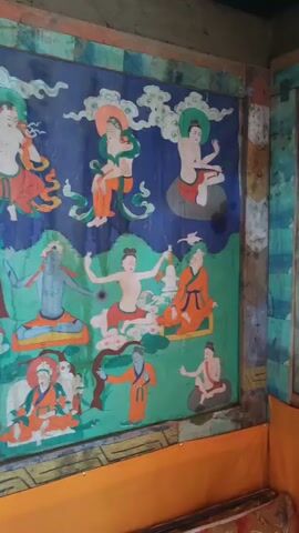 The video shows a mural painted on a wall. The mural depicts several figures in various poses, some seated and some standing. The figures are rendered in a colorful, stylized manner, with distinct facial features and traditional attire. The background is a vibrant blue, with green mountainous landscape elements. The mural is framed by wooden beams at the top and a yellow border at the bottom. The lighting suggests an indoor setting, possibly a temple or historical building, with the wall itself appearing to be made of plaster or a similar material. The painting style and iconography suggest a Buddhist or similar religious theme, likely from East Asian or Himalayan art traditions.
