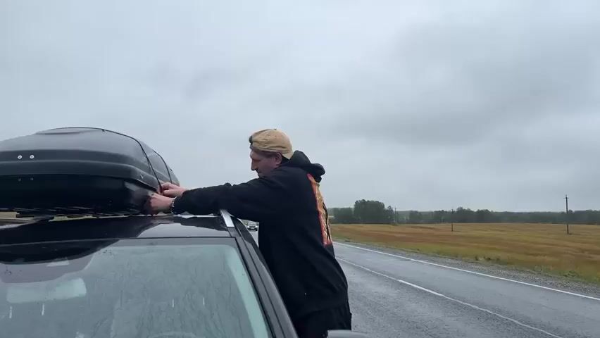A person wearing a black hoodie and a tan baseball cap is standing next to a black car with a black cargo box on its roof. They are reaching up to the cargo box and appear to be adjusting something on it. The car is parked on the side of a wet asphalt road. To the right of the car, a white semi-trailer truck is driving by. In the background, there are fields with yellow and brown vegetation, and the sky is overcast with clouds. Another car, dark blue, drives past the truck.