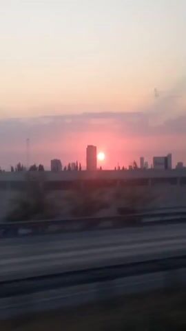 A time-lapse video shows a sunset over a city skyline. The sun is a bright red orb peeking through pink and purple clouds. In the foreground, a blurred car drives by on a highway. Tall buildings and a power line tower are visible against the colorful sky. The scene is captured from a moving vehicle, indicated by the motion blur.
