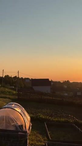 A time-lapse video shows a sunrise over a rural landscape. The sky changes from dark blue to orange and then to a bright yellow as the sun rises. Below, houses and a fenced garden are visible, with two greenhouses in the foreground. The sun, initially hidden, gradually appears as a bright, glowing orb above the horizon.