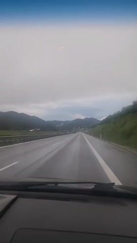 A first-person perspective video captured from inside a car shows a highway stretching into the distance under a cloudy sky. The road appears wet, suggesting recent rain. Mountains covered in green vegetation flank the highway on both sides. In the distance, traffic lights are visible, and signs indicate highway exits or information. The car's dashboard is partially visible in the foreground. A small red light or reflection is present in the sky. The video is short and shows continuous forward movement along the highway.