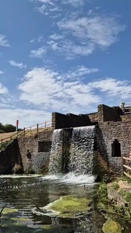 A vertical video shows a waterfall cascading down a stone structure into a body of water. The water in the foreground is green with algae. A wooden fence lines a grassy slope to the left of the waterfall, and another wooden fence is visible to the right. In the distance, a person can be seen walking on a path to the right of the waterfall. The sky is blue with white clouds.