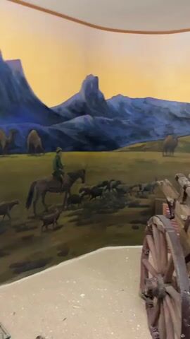 The video pans across a diorama depicting a desert landscape with a mural on the wall. In the foreground, a wooden cart with large spoked wheels is laden with sacks, rugs, and rope. The mural behind the cart shows a shepherd on horseback herding a flock of sheep, camels grazing in the distance, and a yurt with people gathered outside. The landscape in the mural features rolling hills and a jagged mountain peak under a yellow and blue sky.