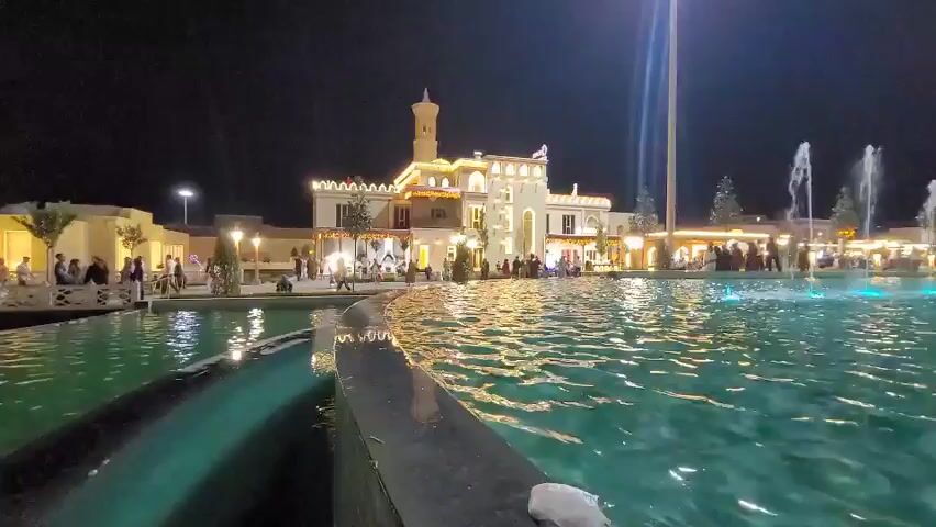 The media file shows a nighttime scene in Satkak, Uzbekistan. The focal point is a large, illuminated building with a tower, resembling a palace or a grand structure. In front of it, a large, shallow pool or plaza features several active water fountains, with some illuminated in blue. The water's surface reflects the lights from the building and surrounding areas, creating a shimmering effect.

There are many people gathered around the building and the fountain area, suggesting a public space, park, or entertainment venue. Some individuals are seen walking, while others appear to be standing and observing the fountains. The overall atmosphere seems lively and festive, with the bright lights and the presence of a crowd.

The time of day is clearly night, indicated by the dark sky and the artificial lighting. The weather appears calm, as the water in the fountains is being projected upwards without being significantly disturbed. The architecture of the building, with its decorative elements and tower, hints at a possible cultural or historical significance to the location. The presence of a flag on top of the building suggests it's a public or official site. There is no discernible text visible in the media file.