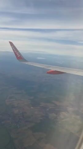 An airplane wing is visible extending from the right side of the frame, with the "easyJet" logo on its surface. The wing casts a shadow over a landscape below. The landscape appears to be rural, with a patchwork of fields and some scattered buildings. The sky above is blue with white, wispy clouds. The perspective suggests the video was taken from inside the airplane, looking out the window.