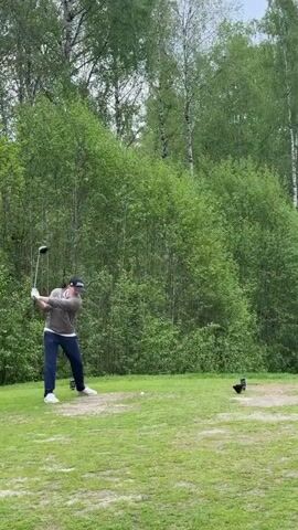 The video begins with a medium shot of a man swinging a golf club on a green golf course. He is wearing a light-colored top, navy pants, white shoes, and a dark baseball cap. Trees line the background. The camera follows the trajectory of the golf ball, panning to a wide shot of the golf course. The course is a green valley surrounded by trees and hills under a cloudy sky. In the distance, a body of water can be seen. The camera slowly pans out, revealing more of the course and the surrounding landscape.