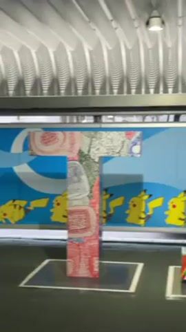 The video shows a series of large letter sculptures and artwork featuring Pokemon characters. The sculptures spell out "TOKYO," each letter having a unique and colorful design. The artwork behind the letters depicts Pokemon characters against a light blue background with snowflake patterns. The scene appears to be inside a train station with a corrugated metal ceiling.