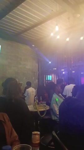 The video shows the interior of what appears to be a bar or club. Multiple people are seated at small tables, drinking and conversing. Some are partially visible from the back or side. The room has a modern, industrial aesthetic with exposed ceilings and metal lattice work. Lighting includes hanging bulbs and colored laser projections, creating a vibrant atmosphere. Drinks are visible on the tables, including beer and soda bottles. The camera pans slightly, showing more of the crowded interior.