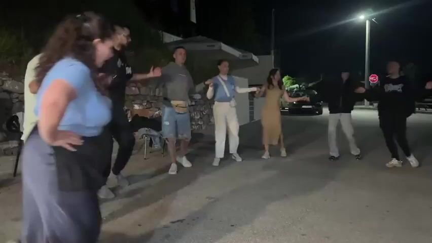 At night, a group of approximately ten people are line dancing in a paved outdoor area. A streetlight illuminates the scene. Some are dancing at the back and some at the front. At the front, they are holding hands. A man in a light green t-shirt with text on the back is the central figure of the front group. Behind him to the left is a man in a black tracksuit. To the right is a woman in a light blue shirt and dark blue skirt. Some chairs are stacked to the left. A white car is parked behind the group dancing in the back.