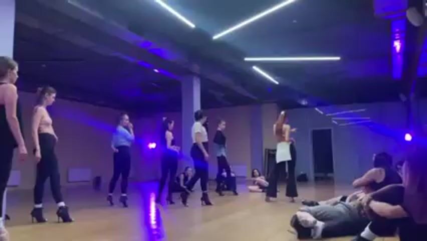 A dance studio is illuminated with a mix of purple and white light, featuring a hardwood floor. A group of about a dozen young women are dressed in various dance attire, including leggings, crop tops, and heels. Some are standing and observing, while others are seated or lying on the floor. One woman, dressed in a white top, white skirt, and heels, stands facing the group, gesturing with her arm as if providing instruction. The environment includes a mirrored wall, a pillar, and a window. The atmosphere suggests a dance rehearsal or class.