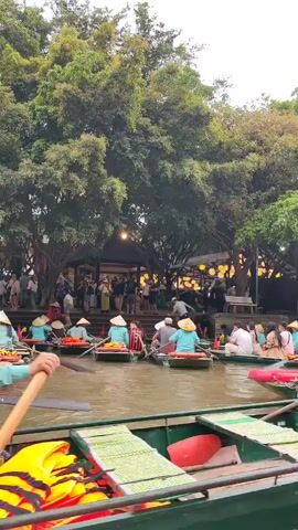 The video shows a waterway crowded with small boats being rowed by individuals wearing conical hats. Many passengers are visible in each boat. The boats are passing in front of a covered area with people milling around and a backdrop of lush green trees. Life jackets and buoys are visible in the boats, suggesting a recreational or tourist activity. The number 1188 is visible on the side of one boat. The sky appears overcast.