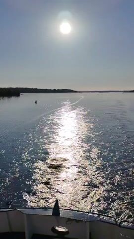 The video, filmed from the stern of a moving vessel, depicts a wide body of water under a bright, sunny sky. The sun, a luminous orb, is high in the center of the frame, casting a brilliant reflection that shimmers along the boat's wake, which extends directly from the foreground into the distance. The water appears calm and blue, with ripples generated by the boat's movement. Distant landmasses, possibly coastlines or islands, are visible on both the left and right sides of the water, adorned with trees. A single dark buoy floats in the water to the left of the wake. The white structure of the boat's stern, including a railing and a dark object on its surface, is visible in the lower portion of the frame. The video maintains a static, eye-level perspective from the moving boat.