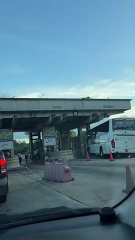 The video shows a toll plaza under a cloudy sky. The perspective appears to be from inside a vehicle, looking forward. In the foreground, a section of a car's interior is visible, including the dashboard. Several lanes of traffic are present. To the left, a red SUV is partially visible. In the center, a pink plastic barrier is situated on the road. Beyond the barrier are toll booths, with several individuals visible, some appearing to be toll collectors or attendants, and others possibly drivers or passengers. One lane has a white bus stopped, with an orange traffic cone nearby. Another lane shows people interacting at a toll booth. The toll plaza structure is concrete, with multiple pillars supporting an overhead walkway or sign area. Signs with text and symbols, some in a foreign language, are attached to the structure. Trees and greenery are visible in the background, suggesting an outdoor environment. The overall impression is of a busy transportation point.