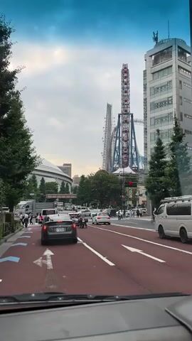 A first-person perspective from inside a car shows a city street with moderate traffic. The road ahead is paved with red asphalt and marked with white lane lines and blue directional arrows. Several cars are stopped in traffic, including a dark gray sedan in the foreground, which is the primary focus. In the distance, a tall Ferris wheel with red gondolas and a roller coaster structure are visible against a cloudy blue sky. Tall buildings and lush green trees line the street. Pedestrians are seen walking on the sidewalk. The overall impression is a bustling urban environment.