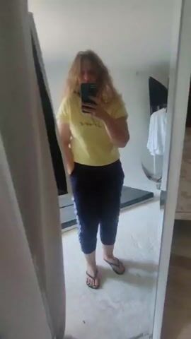 A woman stands in front of a mirror, taking a selfie. She has blonde hair and is wearing a yellow t-shirt and blue athletic pants. She is wearing flip-flops. The mirror is in a room that appears to be a bedroom or dressing area. In the reflection, a bed can be seen, as well as a closet filled with clothes. The room is lit and appears to be messy, with some clutter on the floor.