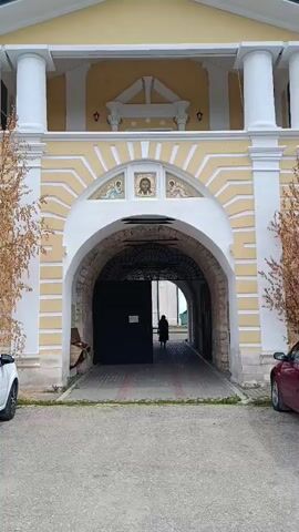 The video shows a view of a yellow and white church belfry with an arched entryway. The camera is positioned in front of the entryway, looking upwards and slightly to the right. The entryway is partially obscured by a dark door. A person in dark clothing is walking through the entryway into the courtyard beyond. The sky is cloudy with patches of blue. Trees with autumn foliage are visible on either side of the entryway. There are cars parked on either side of the entryway.