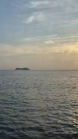 The video shows a seascape. The water is calm, with gentle ripples reflecting the sunlight. In the distance, there is a small island. The sky is a mix of light blue and grey, with the sun setting on the horizon, casting a golden glow over the water.  The viewpoint appears to be from a moving boat.
