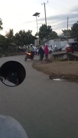 The video, taken from the perspective of someone riding a motorcycle, shows a road in an area that looks like a small town or village. Another motorcycle passes by, and then a red auto rickshaw (tuk-tuk) comes into view and drives past. Several people are walking on the side of the road, one wearing a pink hooded garment. The sky is overcast, and it appears to be either early morning or late afternoon. A solar-powered street lamp is visible along with utility poles and trees.  The video ends with the motorcycle continuing down the road.