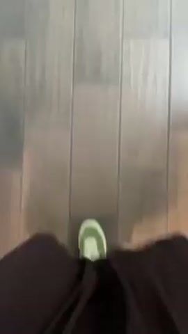 The video shows a first-person perspective of someone walking on a floor made of light-colored, rectangular tiles. The person is wearing dark brown, loose-fitting pants and light green and white sneakers. The camera is shaky due to the movement of walking.