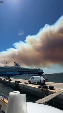 The video shows a harbor scene with a large cruise ship docked in the background. In the foreground, a white van with its rear doors open is parked on a pier. A large plume of smoke is visible in the sky, originating from a fire in the distance. The smoke is a mix of brown and white, suggesting it is a wildfire. The sky is blue and clear, indicating a sunny day. The water is calm and the pier is made of concrete with yellow lines painted on it. There are also some boats and other equipment visible on the pier. The smoke plume is massive and appears to be spreading rapidly. The overall impression is one of a natural disaster occurring in the vicinity.