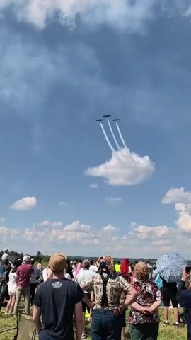 A low-angle shot captures a group of people watching three planes perform an aerial maneuver against a cloudy blue sky. The planes are flying in formation, leaving white smoke trails that curve downwards. The people in the foreground are mostly facing away from the camera, looking up at the sky. They are dressed in casual summer clothing. The environment appears to be an outdoor event, possibly an airshow, with grassy terrain visible.