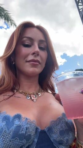A woman with auburn hair, wearing a blue lace-trimmed top and multiple gold necklaces, is drinking a pink beverage from a clear plastic cup with ice. She is outdoors, as evidenced by palm trees and a cloudy blue sky visible in the background. A metal structure is also seen. The woman looks directly at the camera, pausing between sips. Her fingernails are decorated with colorful designs.
