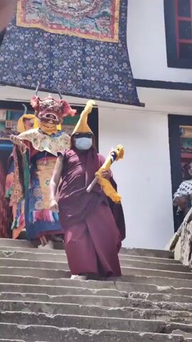 In the video, two individuals are wearing elaborate masks and costumes, possibly for a religious or cultural festival. They are accompanied by a third person wearing a maroon robe, a yellow hat, and a blue face mask, holding a yellow and blue object. They are walking down a set of stone stairs. In the background, a building with traditional architecture and colorful banners is visible. Another person in a white and black patterned outfit is partially visible on the right side of the frame.